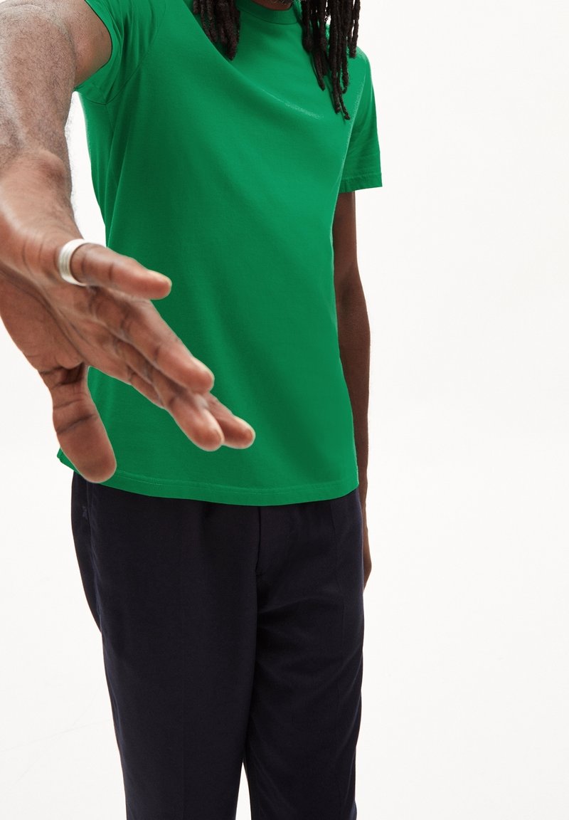 Zelené bavlněné tričko s kulatým výstřihem, krátkými rukávy a rovním lemem. Námořnické kalhoty s jemnou texturou. Ruka se natahuje.