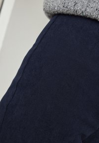 Pantalon en velours côtelé bleu marine avec une texture côtelée, comportant une couture latérale et associé à un pull en tricot gris, présentant un revers plié.
