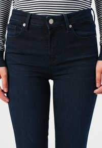 Mørkeblå skinny jeans med knappelinning, frontlommer og en glatt tekstur. Har et klassisk jeansdesign uten utsmykking.
