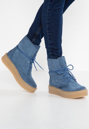 Blue suede ankle boots with thick tan rubber soles worn with dark blue skinny jeans on white background.