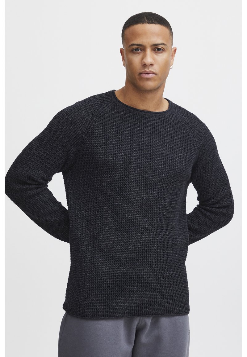 Pull noir en tricot avec un motif texturé, col rond et manches longues, associé à un pantalon gris.