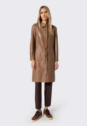 Donna bionda che indossa un lungo cappotto marrone in pelle con bottoni, pantaloni scuri alla caviglia e sneakers con stampa leopardata, in piedi contro uno sfondo neutro.