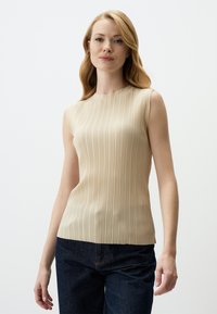 Top beige senza maniche con pieghe verticali, colletto alto e texture liscia, abbinato a pantaloni in denim blu scuro.