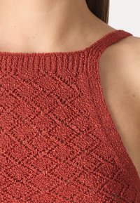 Haut en tricot rouge avec un motif texturé en diamant, des fines bretelles et un col côtelé. Le tissu est doux et légèrement brillant.