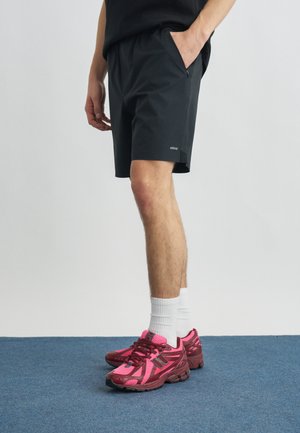 Άτομο που φοράει μαύρη βερμούδα με τσέπη με φερμουάρ και ροζ αθλητικά παπούτσια New Balance, στέκεται σε μπλε χαλί.