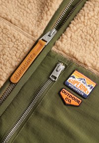 Zielono-beżowa polarowa kurtka z podwójnym zamkiem błyskawicznym, skórzaną zawieszką oraz dwoma haftowanymi naszywkami o tematyce górskiej i outdoorowej.