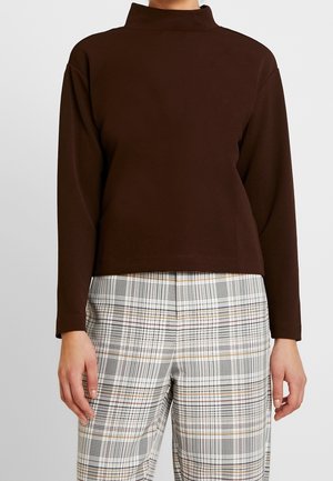 Femme portant un haut marron à manches longues et col montant, et un pantalon blanc à carreaux avec des lignes noires et beiges, debout les bras détendus le long du corps.