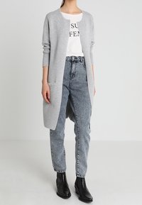 Šedý kardigan s žebrovanou texturou, otevřený vpředu, s dlouhými rukávy, doplněný bílým tričkem s potiskem a vybledlými džíny s vysokým pasem. Černé kotníkové boty dokreslují celkový vzhled.