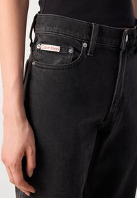 Nærbilde av en person som har på seg høyt midjede svarte Calvin Klein-bukser med rød logo på frontlommen, hånden hvilende ved siden av buksene.