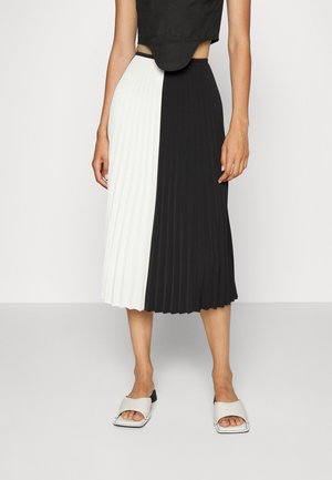 Mujer con falda midi plisada verticalmente en blanco y negro, top corto negro y sandalias con tacón y punta abierta blancas.