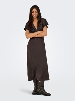 Woman with long dark hair wearing a black short-sleeve midi dress and black leather boots, standing with arms crossed and smiling.