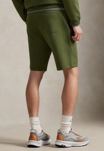 Ελιάς σορτς φερμελής με επίπεδη μέση και πίσω τσέπη, σε συνδυασμό με γκρι αθλητικά παπούτσια και λευκές ριγωτές κάλτσες. Ομαλή υφή.