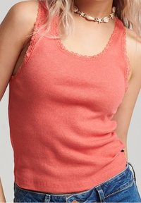 Vrouw in een koraalkleurige tanktop met kantafwerking en een schelpketting, gecombineerd met blauwe denim jeans.