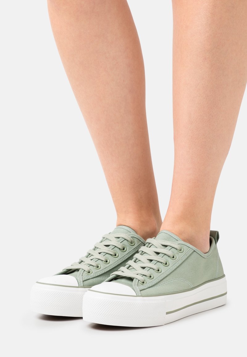 Pieds portant des baskets compensées à lacets de couleur vert clair avec des semelles en caoutchouc blanches et des embouts blancs sur un fond neutre.