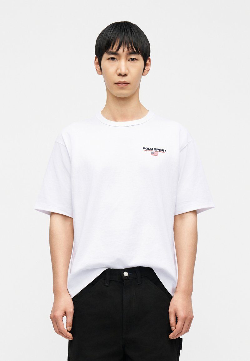 Νέος άνδρας με κοντά μαύρα μαλλιά, που φοράει λευκό μπλουζάκι Polo Sport και μαύρο παντελόνι, στέκεται μπροστά σε απλό φωτεινό φόντο.