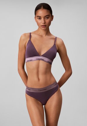 Kvinde iført matchende lilla Calvin Klein bralette og trusser står med hænderne bag ryggen på grå baggrund.