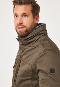 Olivgrüne Jacke mit Stehkragen, ausgestattet mit einem Kunstfellfutter, mit Reißverschlusstaschen und einem strukturierten Stoff. Enthält ein Logo-Patch auf der Schulter.