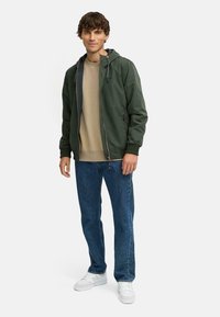 Donkergroen lichtgewicht jack met een capuchon, beige sweatshirt eronder, blauwe spijkerbroek en witte sneakers, gedragen door een staande model.