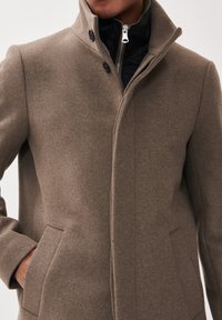 Brauner Wollmischungs-Mantel mit einem hohen Kragen, zwei seitlichen Taschen und schwarer Fütterung, mit einem Reißverschlussverschluss und Knopfdetails.
