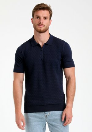 Camisa polo Navy de manga corta, con un patrón de diamantes texturizado, cierre de cremallera y ribete en el dobladillo y las mangas. Usada con jeans azul claro.