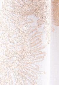 Tessuto ricamato con intricati motivi floreali in beige su uno sfondo bianco, che mostra un aspetto delicato e vissuto.