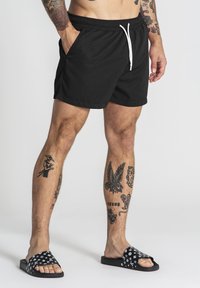 Bañador negro con cintura ajustable, bolsillos laterales y un corte relajado; combinado con sandalias deslizables negras con un patrón, destacando tatuajes en las piernas.