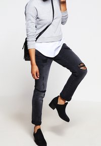 Persona con un suéter gris, camisa blanca, jeans rotos negros, botas negras de tobillo y llevando un bolсe de hombro negro, levantando una pierna.