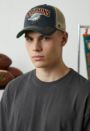 Young man wearing a Dolphins baseball cap and dark gray shirt, with footballs and a Dolphins banner in the background.
