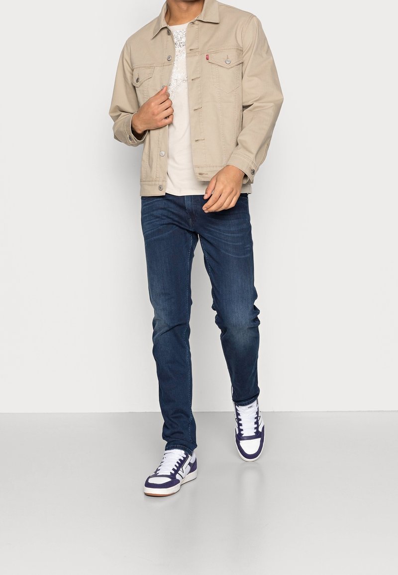 Homme portant une veste beige, un t-shirt blanc avec un motif graphique, un jean bleu foncé et des baskets blanches avec des touches bleu foncé marchant vers l'avant.