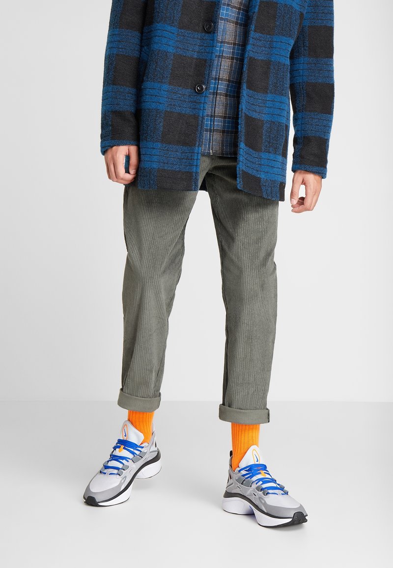 Karierte Jacke in Schwarz und Blau, grau kariertes Hemd, grüne Cordhose und graue Turnschuhe mit blauen Schnürsenkeln und orangen Socken. Lässiges Outfit.