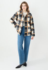 Geruite jas in crèmekleurige, zwarte en oranje tinten; gemaakt van zachte stof met een ritssluiting en een kraag. Combinatie met blauwe jeans.