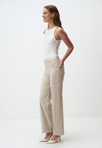 Pantaloni beige a gamba larga con vita alta, progettati con una texture liscia e un dettaglio con bottone in metallo. Abbinati a un top aderente bianco.