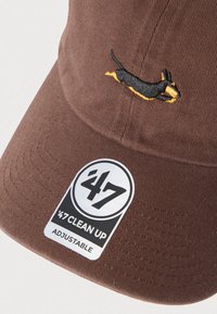 Καφέ καπέλο με μικρό κεντητό σχέδιο σκύλου σε μαύρο και κίτρινο χρώμα. Υφασμάτινη υφή με ρυθμιζόμενο λουράκι και ετικέτα με λογότυπο.