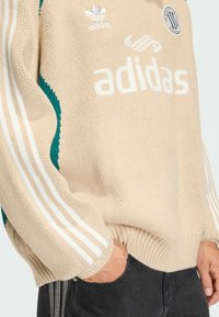 Beżowy sweter dzianinowy z dużym białym logo "adidas", zielonymi akcentami i białymi rękawami z trzema paskami. Ściągacze na mankietach i u dołu; okrągły kołnierz.