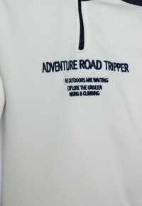 Pull en polaire blanc avec col zippé. Présente des accents bleu marine et un texte brodé inspiré des thèmes et activités de plein air.