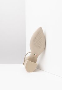 Scarpa stiletto beige con punta affusolata, suola testurizzata e cinturino alla caviglia. Presenta un tacco scolpito e un marchio in rilievo sulla soletta.
