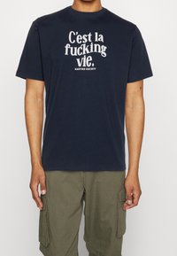 T-shirt en coton bleu marine avec un grand texte blanc qui dit "C'est la fucking vie" et "KAOTIKO SOCIETY" en dessous. Design à col rond.