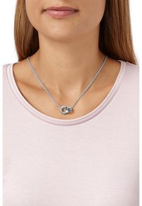 Collana d'argento con design intrecciato e una piccola accentuazione di pietra preziosa, indossata su una maglietta rosa chiaro, posizionata vicino alla clavicola.