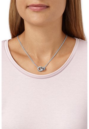 Collana d'argento con design intrecciato e una piccola accentuazione di pietra preziosa, indossata su una maglietta rosa chiaro, posizionata vicino alla clavicola.