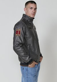 Chaqueta de cuero gris con cuello alto, puños negros contrastantes y un parche bordado rojo en el brazo izquierdo. Bolsillos frontales con cremallera.