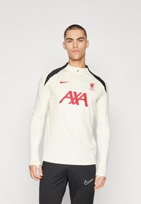 Biała sportowa bluzka z długim rękawem z czarnymi akcentami na ramionach, z czerwonym logo "L.F.C." oraz napisem "AXA". Noszona z ciemnoszarymi spodniami sportowymi, widoczny logo Nike.