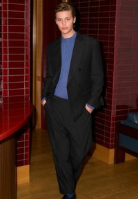 Schwarze gepunktete Anzugjacke über einem blauen Pullover, kombiniert mit gefältelten schwarzen Hosen und schwarzen Schuhen. Der rote geflieste Hintergrund verstärkt den Kontrast.