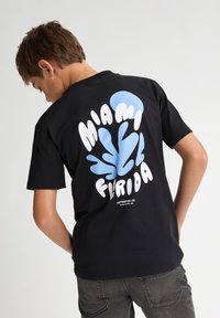 Camiseta negra con un diseño gráfico en la parte posterior que presenta "Miami" y "Florida" en blanco y azul, con una forma orgánica y juguetona.