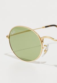 Ochelari de soare rotunzi cu lentile verzi, ramă din metal auriu, accente texturate și suporturi pentru nas transparente. Temele au un design negru și cu model țestoasă.