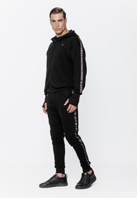 Czarna bluza z kapturem, z zamkiem z przodu i kieszeniami, z długimi rękawami. Połączona z czarnymi spodniami dresowymi, które mają kontrastowe wzory po bokach.