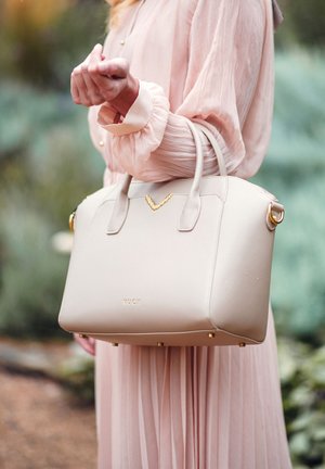 Béžová strukturovaná kabelka vyrobená z hladkého materiálu, s kovovými prvky ve zlaté barvě, šikmým akcentem a čtyřmi zlatými nožičkami na dně.