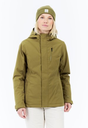 Vrouw in een olijfgroene winterjas met capuchon en bijpassende beanie, staande tegen een witte achtergrond.