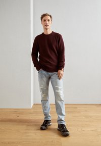 Pull en tricot bordeaux, jeans distressed bleu clair avec déchirures, et baskets noires de sport. Le modèle se tient devant un mur neutre.