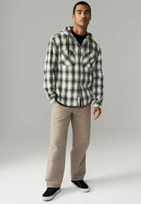 Schwarz-weißer kariert Kapuzenflanellshirt mit zwei vorderen Taschen, kombiniert mit beigen geraden Hosen und schwarzen Turnschuhen mit einer weißen Sohle.