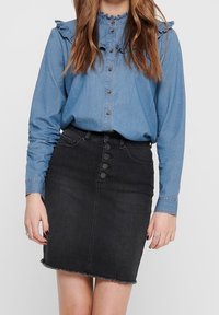 Kvinna iklädd en långärmad blå denimblus med volangkrage och en högmidjad svart denimkjol med knapprad och fransad fåll.